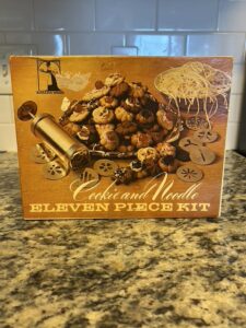 Vintage "Baker's Ware" eleven-piece kit box featuring images of cookies and noodles, a gold-toned cookie press, and various design plates.