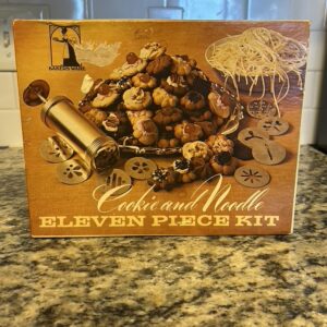 Vintage "Baker's Ware" eleven-piece kit box featuring images of cookies and noodles, a gold-toned cookie press, and various design plates.