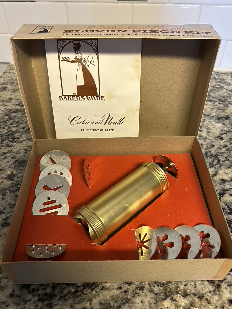Vintage "Baker's Ware" eleven-piece kit box featuring images of cookies and noodles, a gold-toned cookie press, and various design plates.