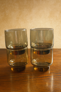 Four stacked, short, smokey grey/brown vintage Arcoroc or Libbey glass tumblers on a wooden surface.
