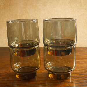 Four stacked, short, smokey grey/brown vintage Arcoroc or Libbey glass tumblers on a wooden surface.