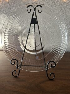 A large, round vintage clear glass serving platter with a ribbed, radiant sunburst design and a plain center, displayed on a metal easel.