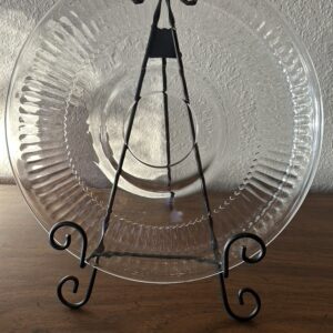 A large, round vintage clear glass serving platter with a ribbed, radiant sunburst design and a plain center, displayed on a metal easel.