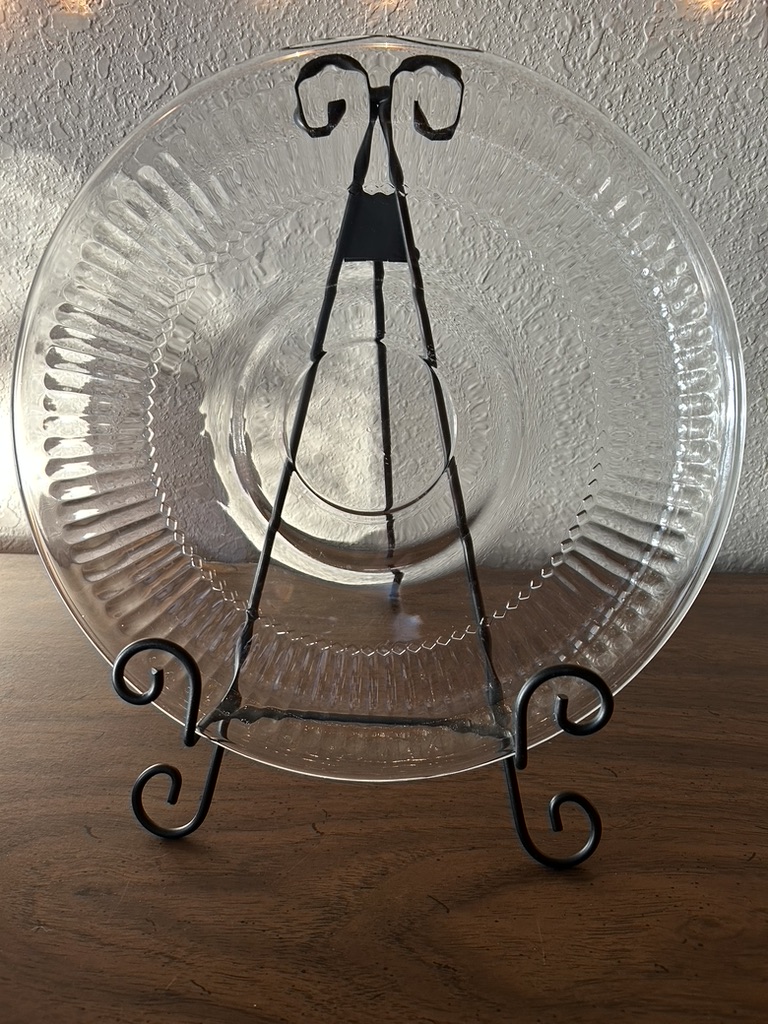 A large, round vintage clear glass serving platter with a ribbed, radiant sunburst design and a plain center, displayed on a metal easel.